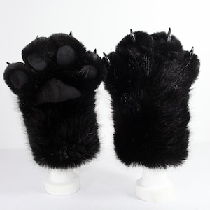 May include: A pair of black faux fur gloves designed as animal paws. One glove displays soft black paw pads, while both feature long, pointed, glossy black claws extending from the fingertips. The plush material covers the entire hand area.
