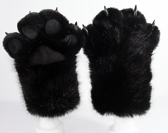 May include: A pair of black faux fur gloves designed as animal paws. One glove displays soft black paw pads, while both feature long, pointed, glossy black claws extending from the fingertips. The plush material covers the entire hand area.