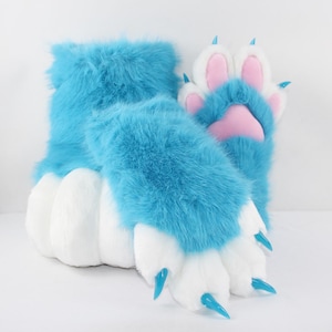 Puede incluir: Un par de patas de peluche de piel sintética azul brillante con puños y dedos blancos. Las patas incluyen almohadillas rosas suaves y garras azules translúcidas y afiladas unidas a cada uno de los cuatro dedos de la pata visible.