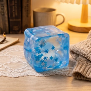 May include: A translucent blue cube-shaped object featuring embedded white and light blue snowflake shapes throughout its structure. The cube has smooth, rounded edges and a glossy finish, displaying a clear, icy appearance with visible depth.