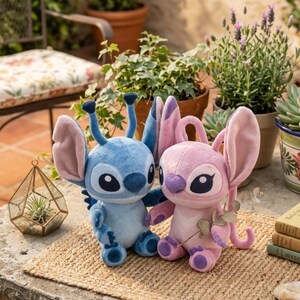 May include: Two plush toys featuring the characters Stitch and Angel. The blue Stitch plush has large ears, antennae, and dark eyes. The pink Angel plush has large ears, a purple nose, and dark eyes. Both are seated upright on a woven mat.