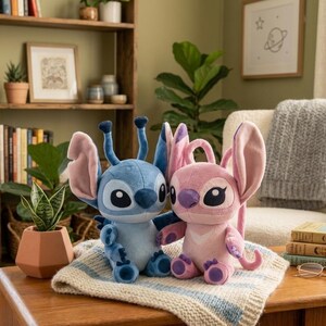 May include: Two plush toys featuring the characters Stitch and Angel. The blue Stitch plush has large ears and antennae, while the pink Angel plush has large ears and a heart-shaped chest pattern. Both sit together on a textured white blanket.