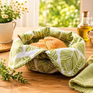 May include: A round fabric bread basket liner featuring a white and green leaf pattern, containing a golden-brown loaf of bread. The fabric is folded over the edges of the basket, displaying a detailed botanical print of stylized leaves.