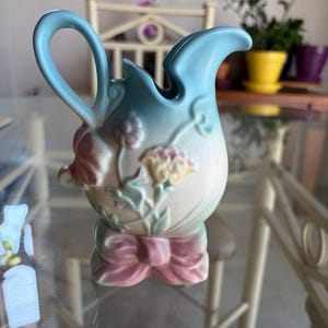 May include: A pastel-colored ceramic pitcher with a light blue handle and spout. The pitcher features floral designs in pink, yellow, and green, and a pink bow at the base. The pitcher is a decorative item.