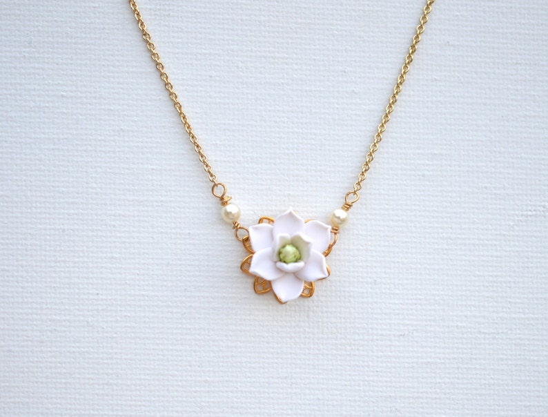 May include: A delicate gold-tone necklace featuring a white flower pendant with a green center. The flower is surrounded by a gold-colored frame and accented with small pearls. The necklace is displayed on a white background.