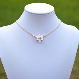 May include: A delicate gold-tone necklace featuring a light pink flower pendant with a small green center. The necklace also includes small white pearl accents. The jewelry is displayed on a white mannequin against a blurred green background.