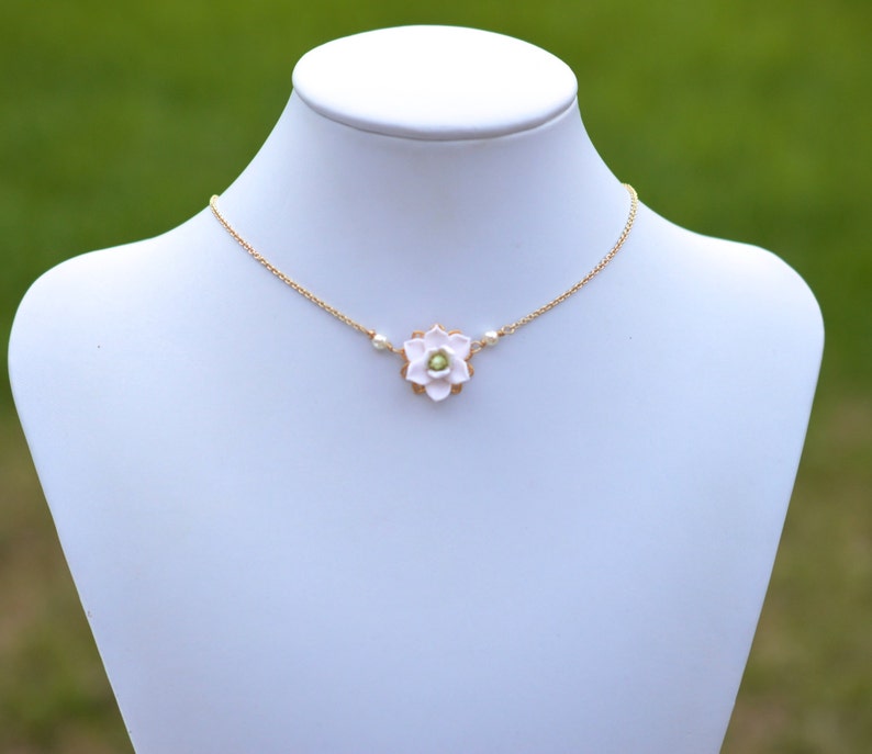 May include: A delicate gold-tone necklace featuring a light pink flower pendant with a small green center. The necklace also includes small white pearl accents. The jewelry is displayed on a white mannequin against a blurred green background.