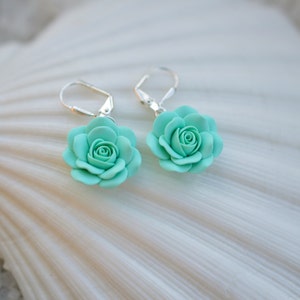 May include: A pair of silver earrings with a light blue rose flower charm. The earrings are displayed on a white seashell.