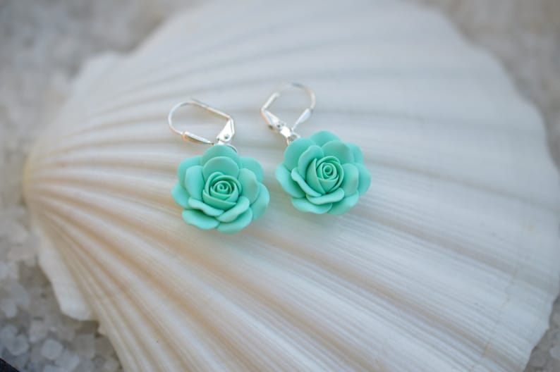 May include: A pair of silver earrings with a light blue rose flower charm. The earrings are displayed on a white seashell.