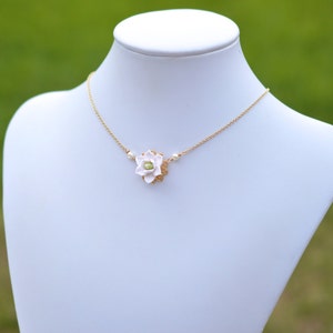 May include: A delicate gold-tone necklace featuring a white flower pendant with a green center. The flower is surrounded by a gold filigree design and accented with small pearls. The necklace is displayed on a white mannequin.