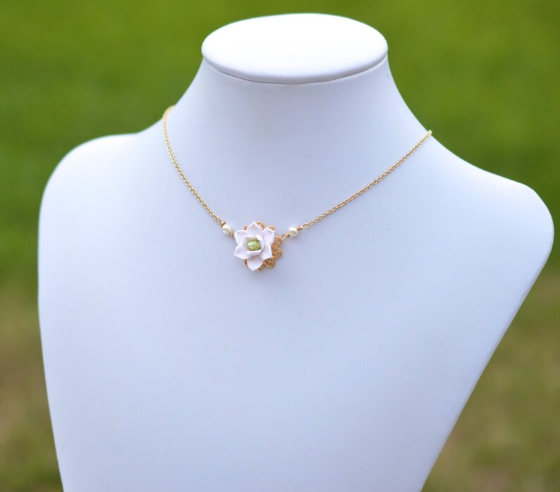 May include: A delicate gold-tone necklace featuring a white flower pendant with a green center. The flower is surrounded by a gold filigree design and accented with small pearls. The necklace is displayed on a white mannequin.