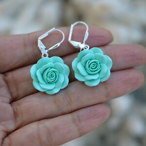 May include: A pair of silver earrings with a light blue rose flower charm. The earrings are dangle style with a hook closure.