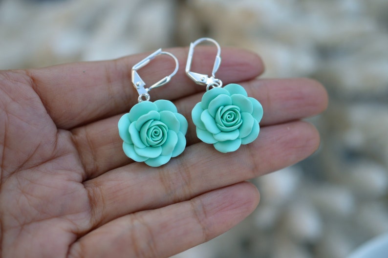 May include: A pair of silver earrings with a light blue rose flower charm. The earrings are dangle style with a hook closure.