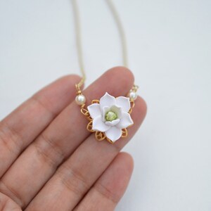 May include: A delicate necklace featuring a white flower pendant with a green center, set in a gold-colored filigree frame. The pendant is accented with small pearls on either side. The necklace hangs on a thin, gold-colored chain.