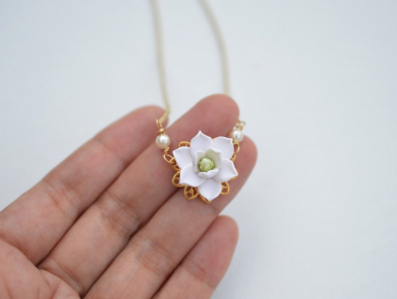 May include: A delicate necklace featuring a white flower pendant with a green center, set in a gold-colored filigree frame. The pendant is accented with small pearls on either side. The necklace hangs on a thin, gold-colored chain.