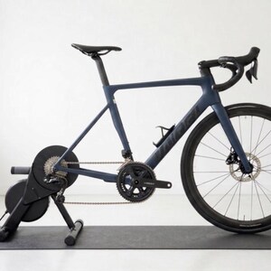 Könnte beinhalten: Ein dunkelblaues Rennrad auf einem schwarzen Trainer, platziert auf einer schwarzen Matte. Das Fahrrad hat schwarze Räder, einen schwarzen Sattel und einen schwarzen Lenker. Der Hintergrund ist eine schlichte weiße Wand.