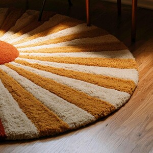 May include: A circular tufted rug featuring a sunburst pattern with alternating rays of mustard yellow and cream white. The center of the rug is a solid burnt orange circle, and the entire piece has a thick, textured pile construction.