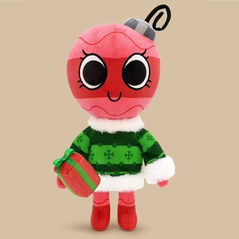 May include: A plush toy shaped like a round pink Christmas ornament with large black eyes, a grey cap, and a black loop on top. It wears a green knitted sweater with white snowflake patterns, white faux fur trim, and red boots, holding a red gift.