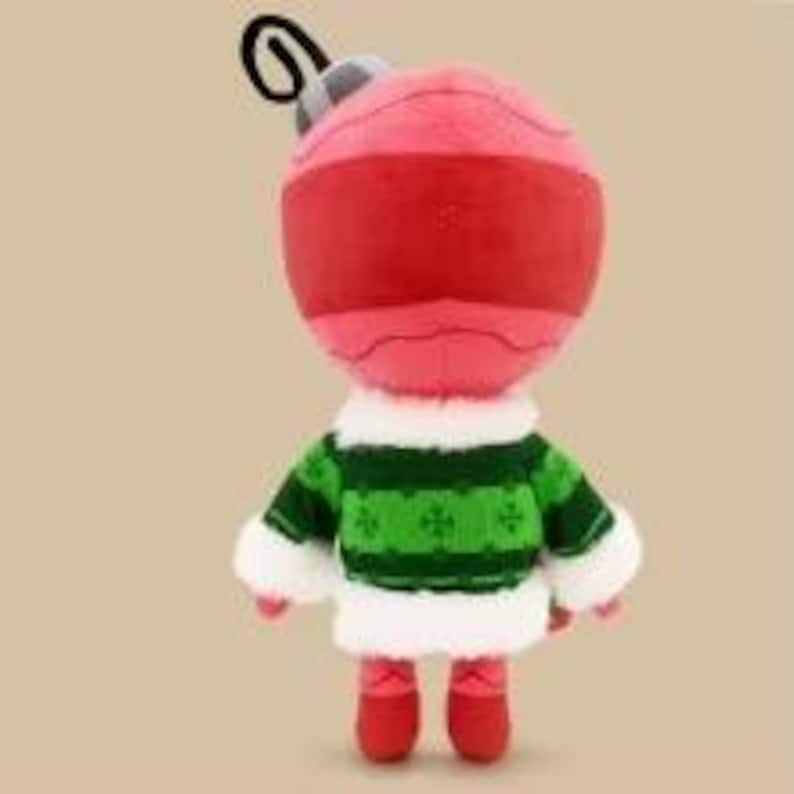 May include: A plush toy shaped like a round pink Christmas ornament with a grey cap and black hanging loop. The toy wears a green sweater featuring a dark green pattern, accented with white faux fur trim at the collar, cuffs, and hem, plus red boots.