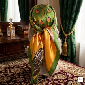 May include: A square silk scarf tied around a head, featuring a vibrant peacock feather pattern in shades of green, orange, and yellow. The design includes detailed eye motifs on the top section and a contrasting golden yellow lower panel.