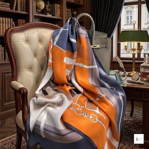 May include: A large square silk scarf featuring a vibrant orange, slate blue, and white color palette. The design displays a detailed graphic print of equestrian-themed leather straps, buckles, and metal hardware elements arranged in a geometric pattern.