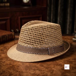 May include: A tan fedora hat featuring an open-weave straw crown and a solid straw brim. The hat is accented with a brown and white striped fabric band that encircles the base of the crown, providing a structured look with a classic silhouette.