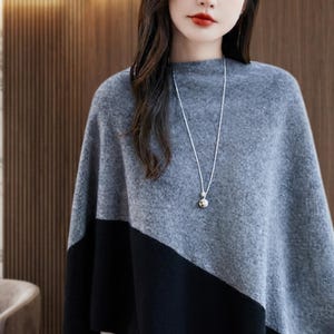 May include: A soft, grey and black color-blocked poncho featuring a diagonal design. The garment has a high neckline and is paired with a long, thin silver-toned chain necklace that features a small, round, metallic pendant at the center.