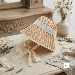 May include: A light brown woven straw sun hat featuring a wide brim and a decorative white lace band around the crown. The hat includes long, matching white lace ribbons attached to the sides, designed to be tied under the chin for secure wear.