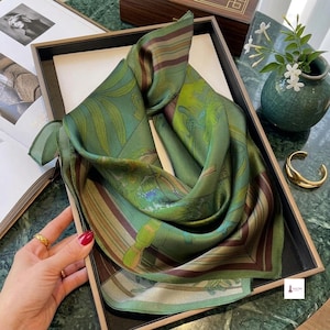 Peut inclure: Un foulard carré en soie présentant une couleur de base vert profond avec des bordures rayées marron et un imprimé botanique détaillé. Le motif comprend des feuilles stylisées et des éléments graphiques subtils dans des tons de vert et marron.