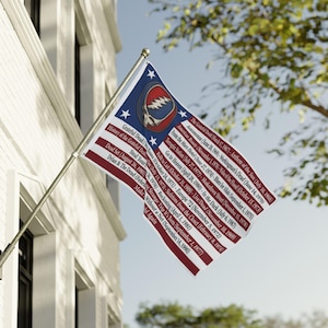 May include: A rectangular flag featuring a blue field with a central Grateful Dead Steal Your Face skull logo and white stars, alongside red and white horizontal stripes printed with a list of album titles and release dates in black text.