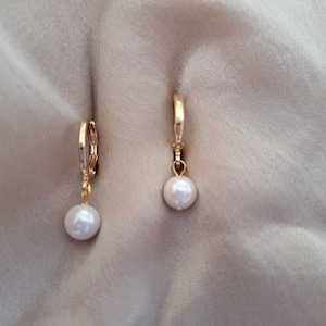 May include: Two gold-coloured leverback earrings, each with a hanging, round, white pearl. The earrings are set against a soft, neutral background, emphasising the jewellery's elegant design. The pearls have a subtle lustre.