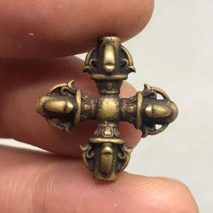 Peut inclure: Un petit pendentif en laiton en forme de croix, composé de quatre branches symétriques et ornées partant d'un centre. Le métal présente une finition dorée antique patinée avec des détails sombres en creux dans les motifs courbes.