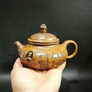 May include: A brown textured clay teapot featuring a small Buddha figure on the lid and a recessed Buddha carving on the side. The body is decorated with black Chinese calligraphy characters. The teapot has a curved handle and a short spout.