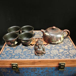 Puede incluir: Un juego de té de arcilla marrón oscuro que incluye una tetera y cuatro tazas a juego, decoradas con motivos florales y paisajes coloridos. Una pequeña figura sonriente se sitúa en el centro sobre una caja de madera con tela floral azul.