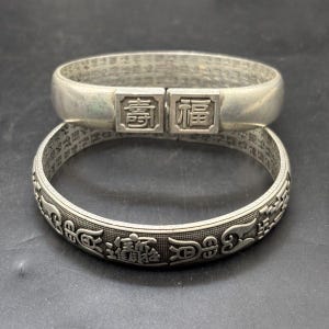 May include: Two silver-toned metal bangles. The top bangle features a smooth band with two square plaques displaying the Chinese characters 壽 and 福. The bottom bangle is decorated with intricate embossed patterns and Chinese characters throughout.