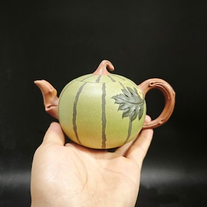May include: A round, light green teapot shaped like a watermelon with dark green vertical stripes. It features a brown spout, a brown handle, and a brown stem-shaped lid knob. A dark green leaf is embossed on the side of the teapot body.