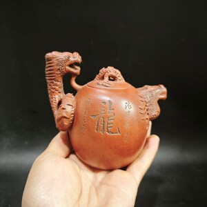 Op de afbeelding: Een roodbruine kleien theepot met gedetailleerde draakvormige handvatten en tuiten. Het bolvormige lichaam is versierd met gegraveerde Chinese karakters en een kleine drakenfiguur op het deksel. Het oppervlak heeft een matte textuur en reliëf.