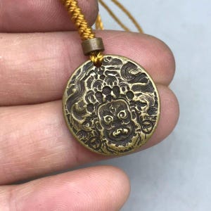 May include: A round brass pendant featuring an embossed, detailed face with a third eye and swirling patterns. The pendant is attached to a thick, braided golden-yellow cord with a small metal bead connector at the top of the circular charm.