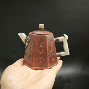 May include: A hexagonal brown teapot with a textured surface featuring vertical columns of engraved Chinese characters. The teapot has a lid with a small knob, a white spout with decorative metal accents, and a white handle with dark grey accents.