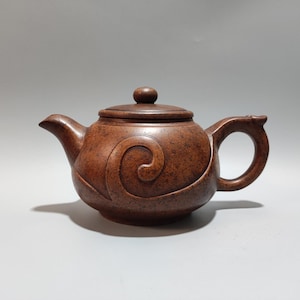 May include: A brown speckled clay teapot featuring a rounded body with a prominent embossed spiral design on the side, a matching lid with a small knob handle, a curved spout, and a sturdy handle extending from the opposite side of the vessel.