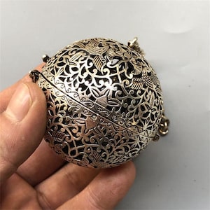 May include: A spherical metal object with an intricate openwork design featuring repeating bird and floral motifs. The surface has a metallic silver finish with dark recessed areas, and a small metal chain is attached to the side of the sphere.