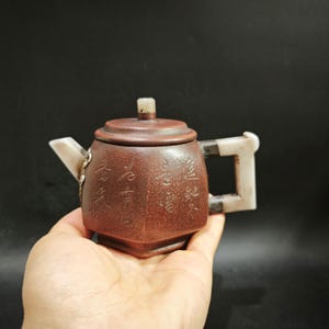 Peut inclure: Une théière hexagonale en argile brune ornée de caractères chinois gravés sur le corps, un couvercle avec un petit bouton, ainsi qu'un bec et une anse en pierre blanche. La théière présente une surface texturée et des accents en pierre claire.