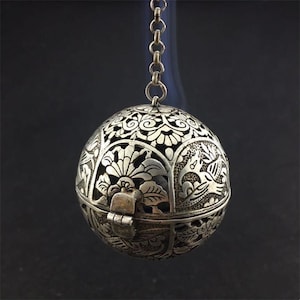 Op de afbeelding: Een bolvormige zilverkleurige metalen pomander met ingewikkelde uitgesneden filigraanpatronen met bloemmotieven en vogelfiguren. Het object hangt aan een korte metalen ketting en bevat een kleine scharnierende sluiting aan de voorzijde.