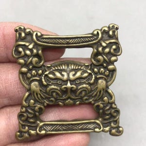 May include: A rectangular brass belt buckle featuring an intricate relief design of a stylised animal face in the centre, surrounded by ornate scrollwork patterns and two horizontal bars with cross-hatched detailing at the top and bottom edges.