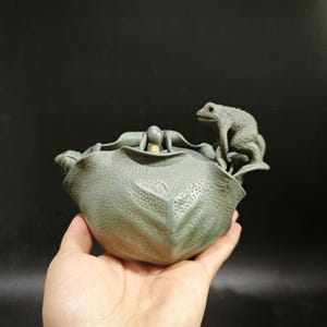 May include: A matte sage green ceramic tea pet shaped like a textured lotus leaf. A small, detailed frog sits on the edge of the leaf, while a tiny, light-coloured duckling head emerges from the centre of the folded leaf structure.