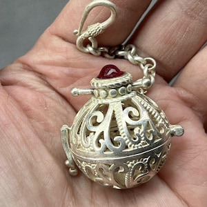 May include: A spherical silver pendant featuring intricate openwork filigree patterns and a smooth, deep red cabochon stone set at the top. The pendant is attached to a short silver chain with a hook clasp, showcasing detailed metalwork.