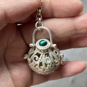 May include: A spherical silver pendant featuring intricate openwork scroll patterns and a small, circular, vibrant green stone set at the top. The pendant is attached to a silver jump ring and chain link, showcasing a detailed metalwork design.
