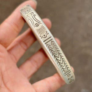 Op de afbeelding: Een zilverkleurige metalen armband met een gegraveerd patroon van geometrische lijnen, chevrons en cirkelvormige motieven. Het oppervlak heeft een matte afwerking met donkere verdiepte delen die de lineaire en cirkelvormige ontwerpen benadrukken.