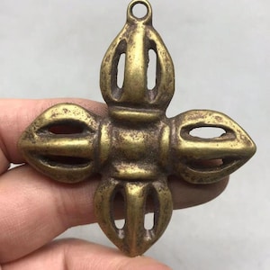 Peut inclure: Un pendentif en métal de couleur laiton en forme de croix, doté d'un moyeu central carré avec quatre branches symétriques ajourées s'étendant vers l'extérieur. L'objet présente une finition texturée et patinée avec une boucle circulaire au sommet.