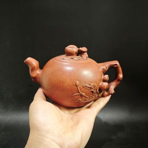 May include: A reddish-brown clay teapot featuring a textured surface with carved botanical details of leaves and stems. The lid is topped with a small, sculpted fruit-shaped finial, and the handle and spout are designed to resemble natural tree branches.